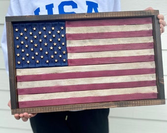 May include: A framed American flag made of repurposed fabric strips. The flag is displayed in a dark wood frame with a distressed finish. The stars are blue and gold, and the stripes are red and white.