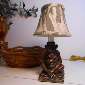 May include: A decorative table lamp featuring a monkey figurine base holding a baby monkey. The lamp has a beige and gray leaf-patterned lampshade. The lamp is on a white surface.