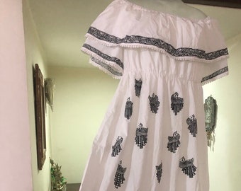 May include: White off-the-shoulder dress with black decorative patterns. The dress features a ruffled neckline and hem, and is adorned with repeating bird-like figures. The dress is floor-length and has a flowing silhouette.