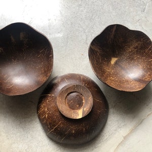 May include: Three brown coconut bowls, two are shallow and one is deeper with a raised centre. The bowls are made from natural coconut shells and have a rustic, organic look.