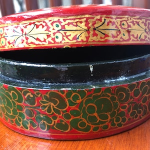 Pode incluir: Uma caixa redonda de papel maché pintada de vermelho, preto e dourado com um desenho floral. A caixa está aberta e a tampa está em cima da caixa.