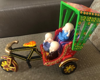 May include: A colorful wooden toy rickshaw with a green canopy and three small figures sitting inside. The rickshaw has a black frame and wheels with yellow and brown details.