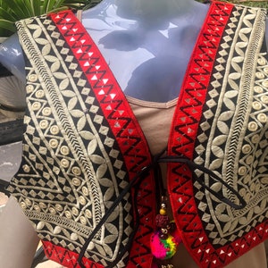 May include: A black and cream embroidered vest with red trim and a tassel tie. The vest has a geometric pattern and is decorated with small mirrors.