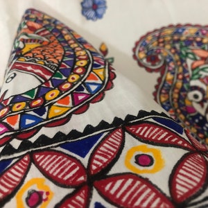 May include: Close-up of a white fabric with colorful hand-painted paisley designs. The fabric features intricate patterns in red, blue, yellow, green, and orange.