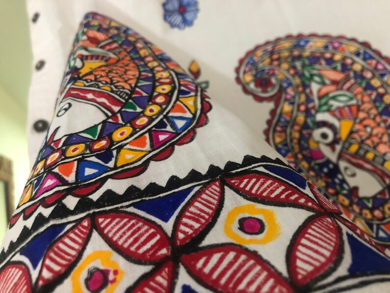 May include: Close-up of a white fabric with colorful hand-painted paisley designs. The fabric features intricate patterns in red, blue, yellow, green, and orange.