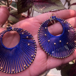 Könnte beinhalten: Ein Paar große Creolen aus blauem Garn und goldenen Perlen. Die Ohrringe sind rund und haben ein fächerartiges Design.