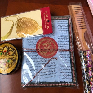 Puede incluir: Un conjunto de regalos de temática india envueltos en plástico transparente. El conjunto incluye una pequeña caja redonda de madera con intrincados diseños en oro y negro, un libro con una cubierta roja y dorada, un peine de madera y un marcapáginas dorado con un borla.