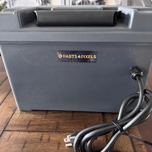 May include: A gray rectangular storage container with a black handle and a power cord. The container has the text "PARTS 4 PIXELS" on the front. The power cord is black and has a standard US plug.