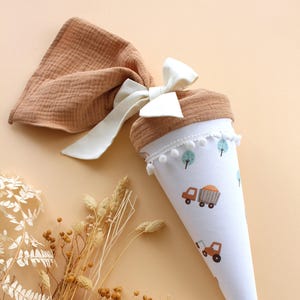 May include: A white and brown school cone with a truck and tree pattern. The cone is decorated with white pom-poms and a white bow. A brown fabric is attached to the top of the cone.