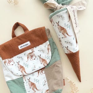 Peut inclure: Un sac à dos en patchwork marron et vert avec un imprimé kangourou et le texte "Little Miss Sophia". Un sac en forme de cône assorti marron et vert avec un imprimé kangourou et le texte "Little Miss Sophia".