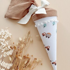 May include: A white and brown cone-shaped gift bag with a truck and tree pattern, trimmed with white pom-poms. A matching brown fabric is tied with a white bow. Dried flowers are in the foreground.