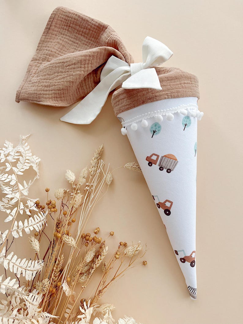 May include: A white and brown cone-shaped gift bag with a truck and tree pattern, trimmed with white pom-poms. A matching brown fabric is tied with a white bow. Dried flowers are in the foreground.