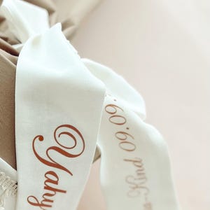 May include: Close-up of white fabric ribbons with brown text. One ribbon reads "Yahya" in a cursive font. The other ribbon reads "Klea-Kind 09.09." in a similar font. The ribbons are tied around a beige fabric.
