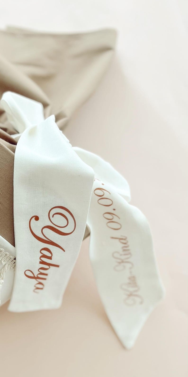 May include: Close-up of white fabric ribbons with brown text. One ribbon reads "Yahya" in a cursive font. The other ribbon reads "Klea-Kind 09.09." in a similar font. The ribbons are tied around a beige fabric.