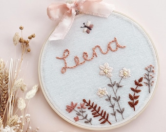 May include: Embroidered hoop art featuring the name "Leena" in copper thread, with floral and butterfly details. A light pink ribbon bow and hanging loop are attached. The artwork is set against a light blue fabric background.