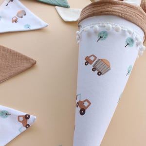 May include: A white fabric cone-shaped school bag with a brown rim and white pom-poms. The bag features a repeating pattern of brown trucks and green trees. Several matching bibs with truck and tree patterns are also visible.