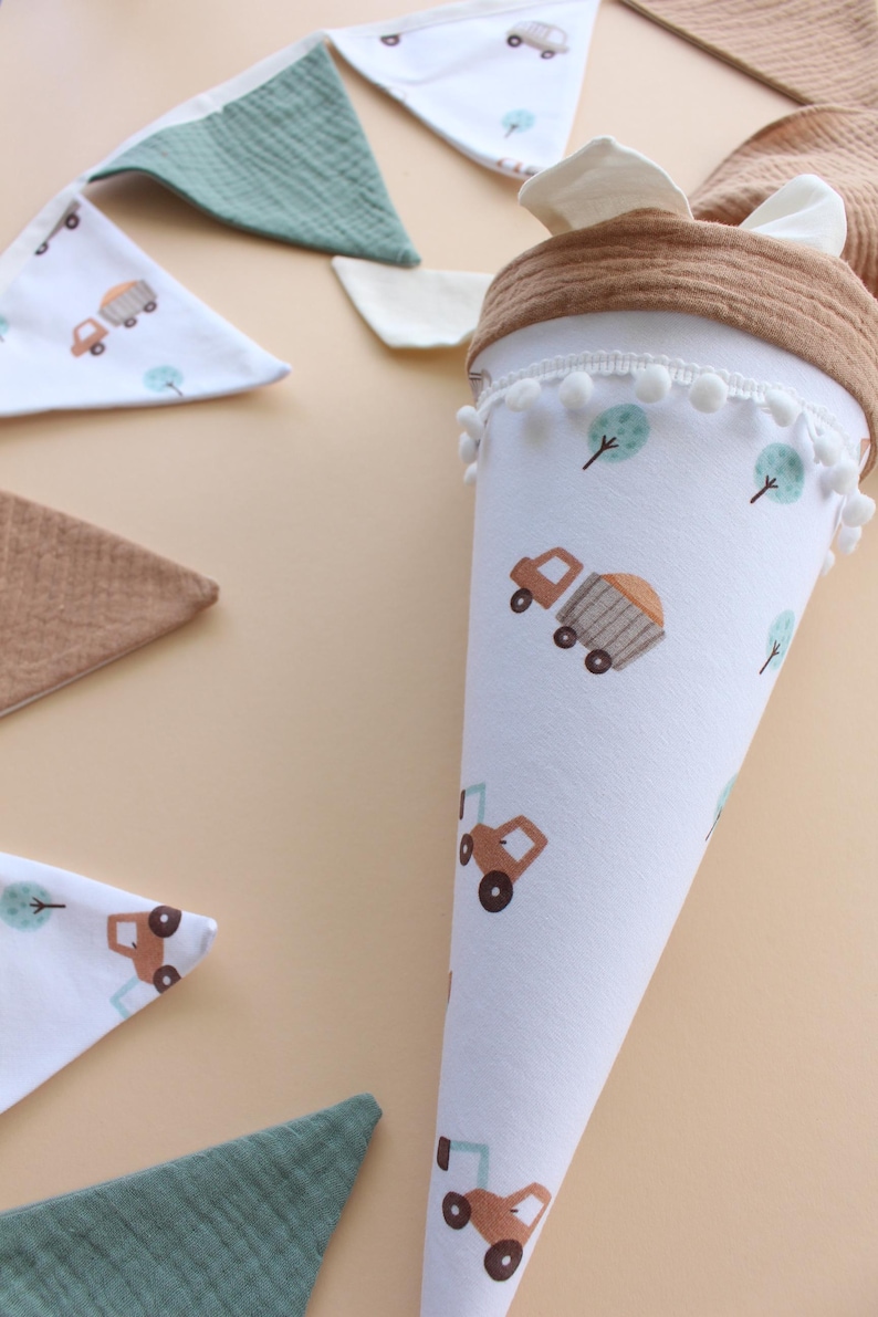 May include: A white fabric cone-shaped school bag with a brown rim and white pom-poms. The bag features a repeating pattern of brown trucks and green trees. Several matching bibs with truck and tree patterns are also visible.