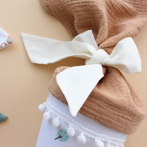 May include: A baby gift set featuring a brown muslin fabric tied with a cream-colored bow. The set includes a white bib with a tree design and pom-pom trim, and a green muslin bib. The background is a light tan color.
