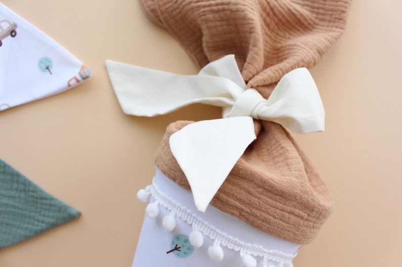 May include: A baby gift set featuring a brown muslin fabric tied with a cream-colored bow. The set includes a white bib with a tree design and pom-pom trim, and a green muslin bib. The background is a light tan color.