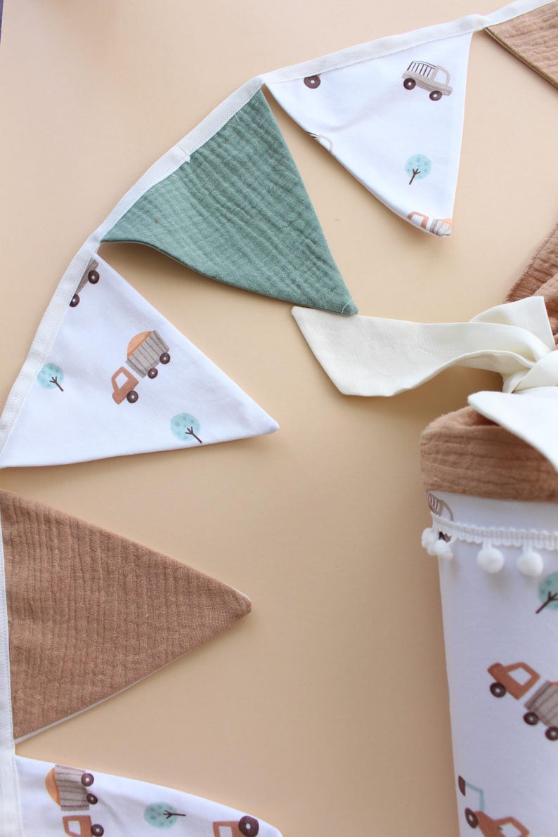 May include: A collection of nursery decor featuring a banner and a container. The banner has alternating triangle flags in white with truck and tree illustrations, green, and brown. The container is white with a truck and tree pattern and a pom-pom trim.