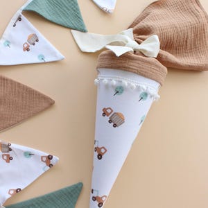 May include: A white, cone-shaped school bag with a brown top and a white bow, decorated with trucks and trees. A matching banner with trucks and green and brown triangles is also visible. The bag is trimmed with white pom-poms.