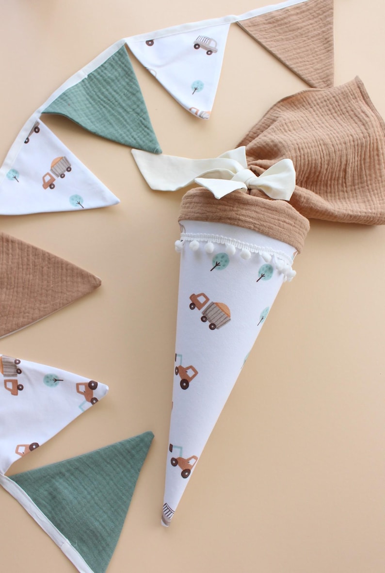 May include: A white, cone-shaped school bag with a brown top and a white bow, decorated with trucks and trees. A matching banner with trucks and green and brown triangles is also visible. The bag is trimmed with white pom-poms.