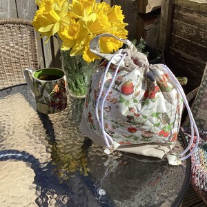 May include: A white drawstring bag with a floral print featuring birds, apples, and other nature-inspired elements. The bag has white drawstrings and is sitting on a glass table with a mug and a vase of yellow daffodils.
