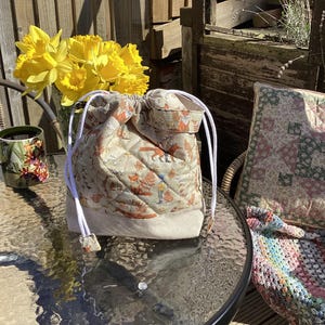 May include: A beige drawstring bag with a fox pattern, white cords, and a quilted design. The bag is on a glass table, alongside a vase of yellow daffodils and a floral mug. The bag's base is beige.