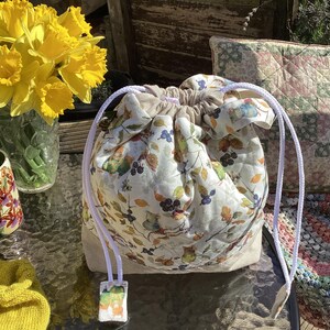 May include: A white drawstring bag with a floral print featuring leaves, berries, and birds. The bag has a white drawstring and is made of a soft, quilted fabric.