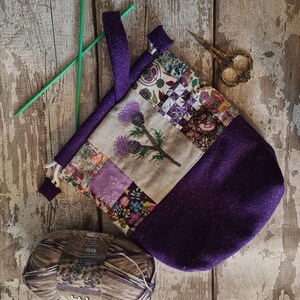 May include: Purple project bag with a patchwork design featuring a floral pattern and a single embroidered thistle. The bag has a drawstring closure and a handle. A skein of brown yarn is in the foreground.