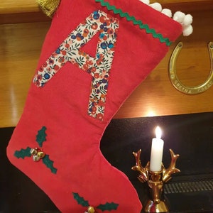 May include: Red Christmas stocking with a white pom-pom trim, green and white trim, and a fabric letter 'A' with a floral pattern. The stocking is hanging on a wooden mantel with a horseshoe and a lit candle.
