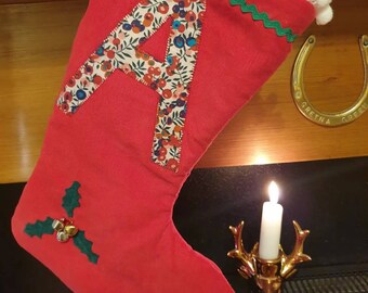 May include: Red Christmas stocking with a white pom-pom trim, green and white trim, and a fabric letter 'A' with a floral pattern. The stocking is hanging on a wooden mantel with a horseshoe and a lit candle.