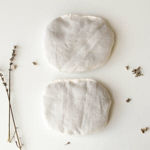 May include: Two white linen eye pillows, each with a simple sewn seam down the center. The pillows are on a white background.