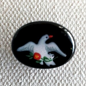 以下が含まれることがあります： 黒い楕円形の背景に、赤い花と緑の葉を持つ白い鳩が描かれています