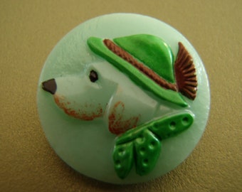 Può includere: Un bottone verde e marrone con un'immagine in rilievo di un cane che indossa un cappello verde. Il cane ha il naso marrone e il pelo bianco.