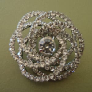 Peut inclure: Broche argentée en forme de fleur, ornée de nombreux petits cristaux transparents. La broche présente un cristal central plus grand, et la forme générale rappelle une rose. Les cristaux scintillent sur un fond vert clair.