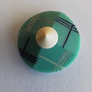 Könnte beinhalten: Ein grüner und weißer Plastikknopf mit geometrischem Muster. Der Knopf hat eine erhabene, weiße, kegelförmige Mitte.