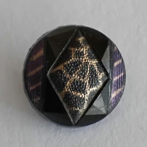 以下が含まれることがあります： 中央に黒と金のテクスチャパターンが施された菱形のインレイがある円形のボタン。周囲の表面は、滑らかな黒いセグメントと、紫と金のストライプが入った布のような素材のセクションが交互に配置されています。