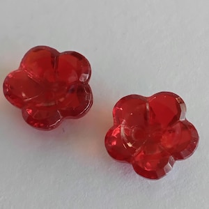 May include: Two translucent red buttons shaped like five-petaled flowers. Each button features a faceted surface that catches the light, creating a glossy appearance. The buttons have a central hole design for attachment to fabric or garments.