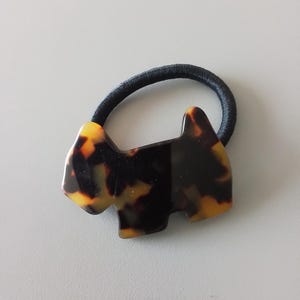 May include: A tortoiseshell-patterned hair tie featuring a stylized dog-shaped charm. The charm is a mix of amber and dark brown hues, attached to a black elastic band. The accessory is designed for hair styling.