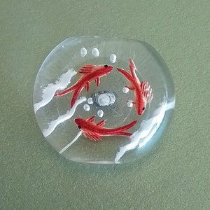 May include: A clear glass paperweight featuring a red and white design of three fish swimming in a circle. The fish are outlined in black and have white accents.