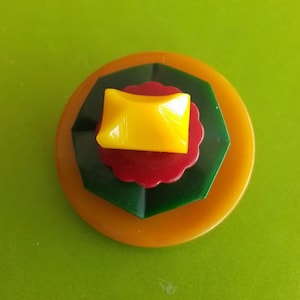 May include: A layered, colorful button with a yellow square top, a red scalloped layer, a green geometric layer, and a large orange circular base. The button is set against a green background.