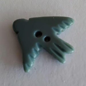 May include: A two-hole button shaped like a bird in flight, featuring a smooth, matte, slate-blue finish. The button has a distinct head, two outstretched wings with molded feather details, and a pointed tail, all rendered in a solid, muted blue color.