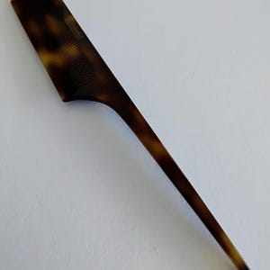 May include: A tortoiseshell-patterned comb with a long, pointed handle. The comb's teeth are visible on the rectangular head, and the handle tapers to a fine point. The color is a mix of brown and amber.