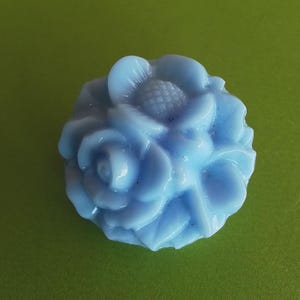 May include: A light blue glass rose flower button. The rose is made of multiple layers of petals and has a textured centre.