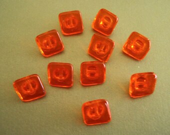 May include: Ten orange plastic buttons with a raised design. The buttons are square shaped and have rounded corners.
