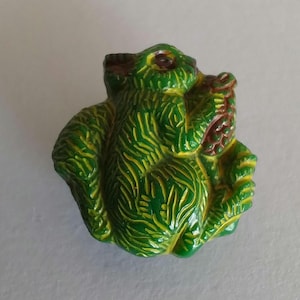 May include: A small, green figurine of a creature with yellow detailing. The figurine has a textured surface and is holding a brown object. The creature's eyes are visible. The figurine is likely a decorative item or collectible.