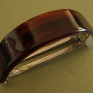 May include: A hair barrette featuring a curved, translucent dark brown tortoiseshell-patterned top piece attached to a silver-toned metal spring-loaded clasp mechanism designed to secure hair in place with a simple snap-shut closure.