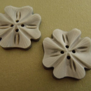May include: Two light grey, flower-shaped buttons with four petals and three holes for sewing. The buttons have a textured surface and are set against a light gold background. Each button measures approximately 2.5 cm in diameter.