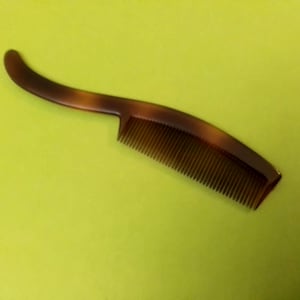 May include: A brown tortoiseshell comb with a curved handle. The comb has fine teeth and a smooth, polished finish. The handle curves gracefully, providing a comfortable grip. The comb is designed for hair grooming.
